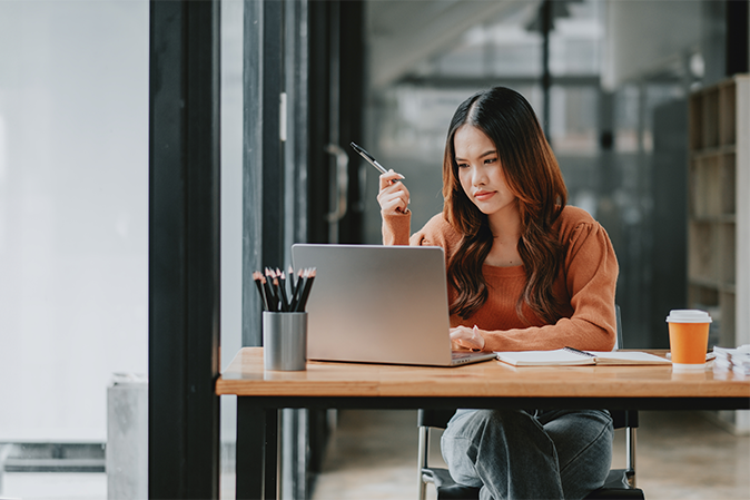 Introspective businesswoman contemplates her work, pen in hand, while sitting at her laptop in a well-organized office space.