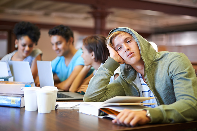Young man burnt out sitting at a table in college classroom with other students