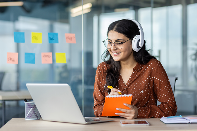 Young woman wearing headphones while watching a video on laptop and taking notes
