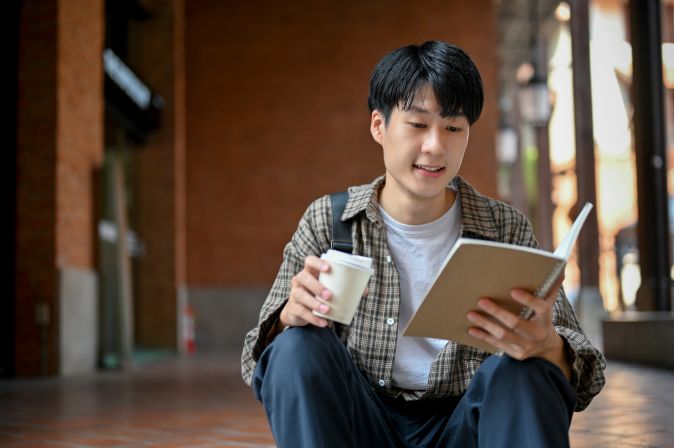 Image of a student sitting and reading a book while holding a cup.