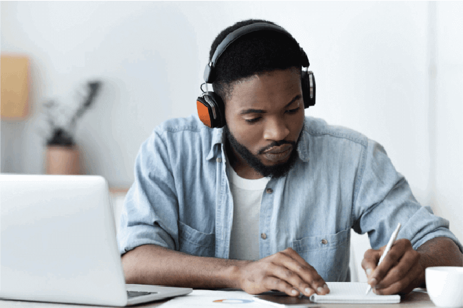 Young man wearing headphones taking notes sitting in front of a laptop