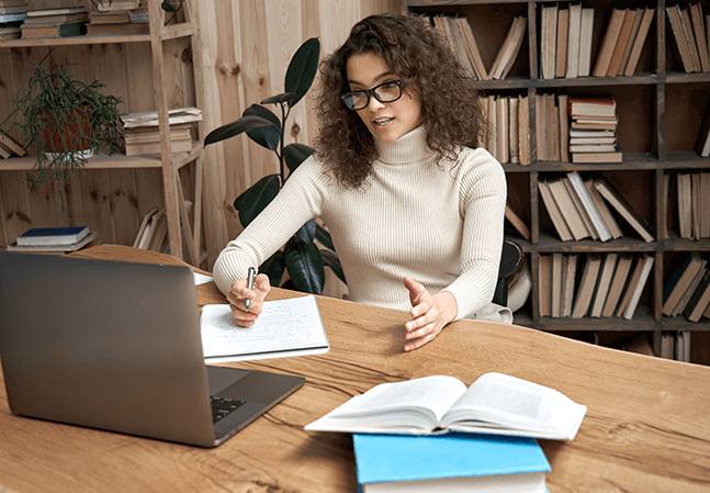 Woman sitting at a desk taking notes in front of a laptop teaching a class online