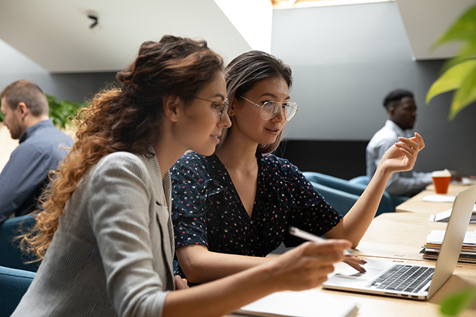 Female asian mentor teacher instructor teaching caucasian businesswoman help intern explaining online project instructing new worker trainee looking at laptop computer write notes sit at office desk