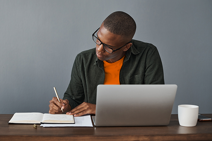 Young man sitting at a desk writing notes in front of a laptop