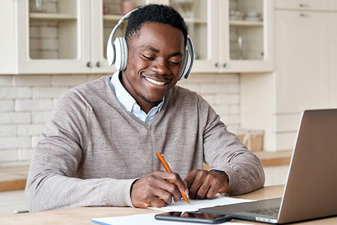 Young man wearing a headset while smiling and taking notes in front of a laptop and mobile phone