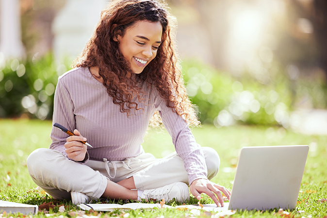 Young woman smiling while studying on a laptop taking notes outside